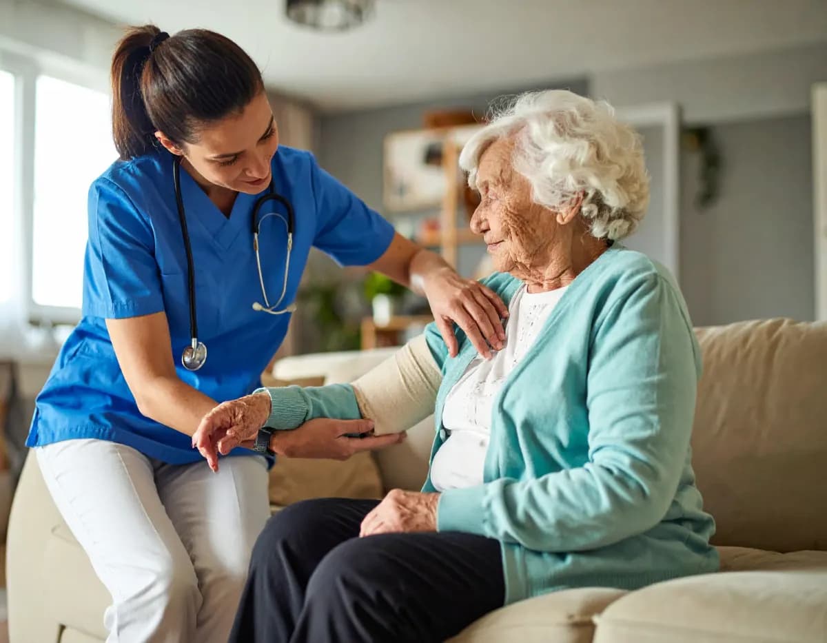 O asistentă medicală o sprijină pe o doamnă în vârstă, acasă, pe canapea